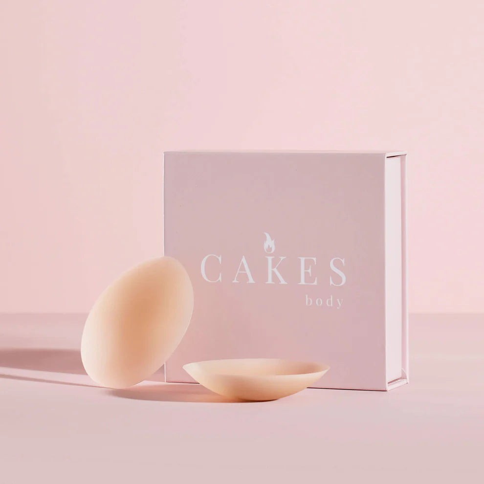 Two silicone breast forms next to a 'CAKES' branded box on a pink background